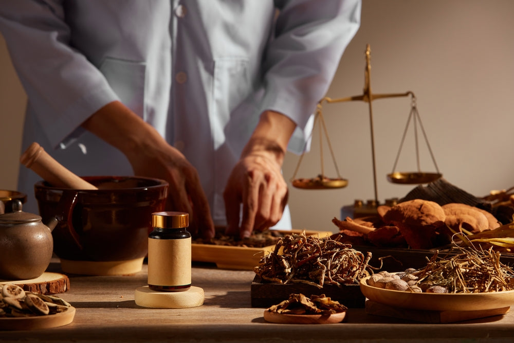 Practitioner preparing traditional herbal medicine with dried roots, mortar, and balance scale on table.
